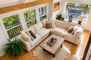 A modern living room with a sectional sofa, a wooden coffee table, and large windows with a view of greenery. - Lakefront, 3 Amazing Decks, Tennis/Pickle Ball, Boat Slip, Hot Tub, Bed Swing
 (Oakland)