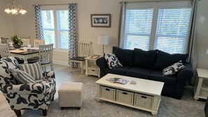 Una sala de estar con un sofá azul oscuro, un sillón estampado y un reposapiés. Hay una mesa de centro con revistas y una lámpara. La habitación tiene una zona de comedor con una mesa y sillas, y ventanas amplias con persianas.