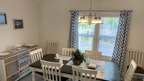 Comedor con mesa de madera oscura, sillas blancas, lámpara colgante y cortinas estampadas.