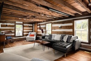A log cabin interior with a wooden ceiling, a black leather sofa, white armchairs, and a rustic dining area. - Walk to Old Rag | Hot Tub | Mountain Farmhouse (Sperryville)