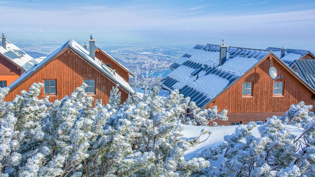 A wooden cabin with a sloped roof, surrounded by snow-covered trees, and a view of a distant landscape.