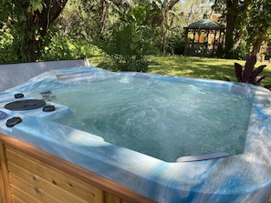 A hot tub with a wooden frame and a transparent cover, surrounded by lush greenery and a gazebo in the background. - Sleeps 20+, 2 houses, Swim-Spa, hot tub, bachelor and party place (Tampa)
