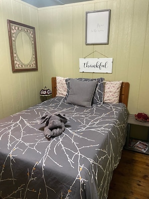 A neatly made bed with a dark grey bedspread featuring a white tree branch pattern, flanked by two pillows with a floral design and a grey pillow. A plush toy is placed on the bed. The room has light green walls, a framed mirror with a floral design, and a 'thankful' sign on the wall. - Charming lake community cottage with hot tub PET FRIENDLY (Glen Spey)