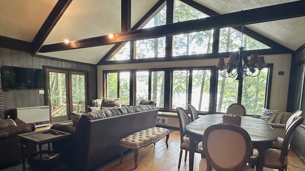 A spacious living room with a large window, a fireplace, and a dining area with wooden furniture. - Private Lake Front Adirondack Retreat on Loon Lake-20 Min from Lake George, Gore (Chestertown)