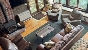Un salon chaleureux avec une cheminée, des canapés en cuir, une table basse et de grandes fenêtres donnant sur l’extérieur.
