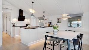 Een moderne keuken met witte kasten, een centraal eiland, hangende lampen en uitzicht op zee door een groot raam.