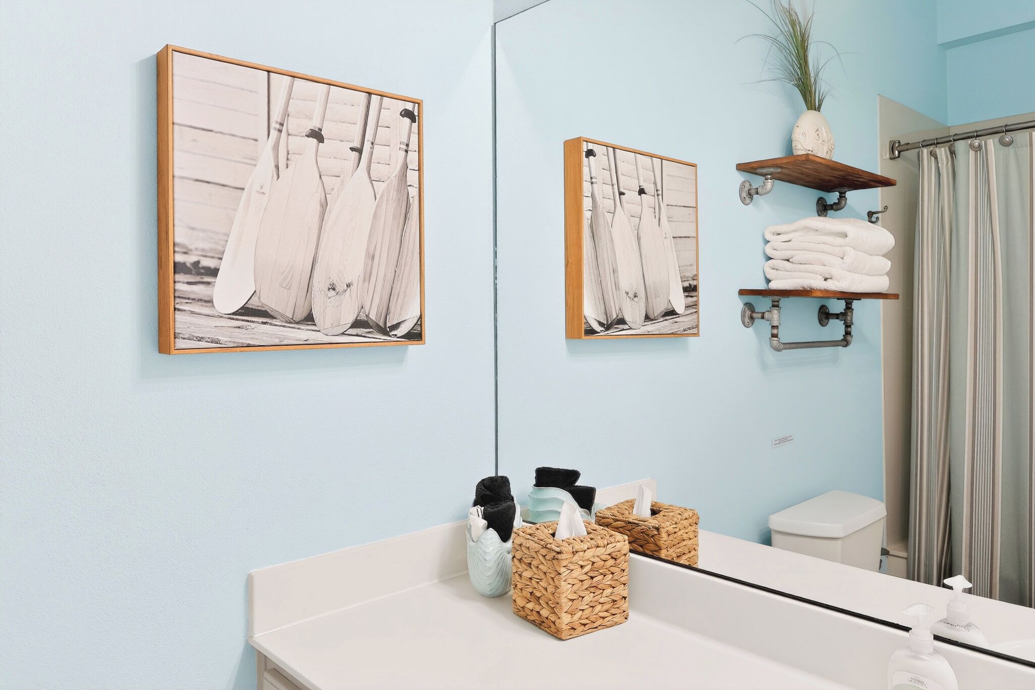 A bathroom with a light blue wall, a wooden shelf with towels, a mirror, and two framed pictures of boats.