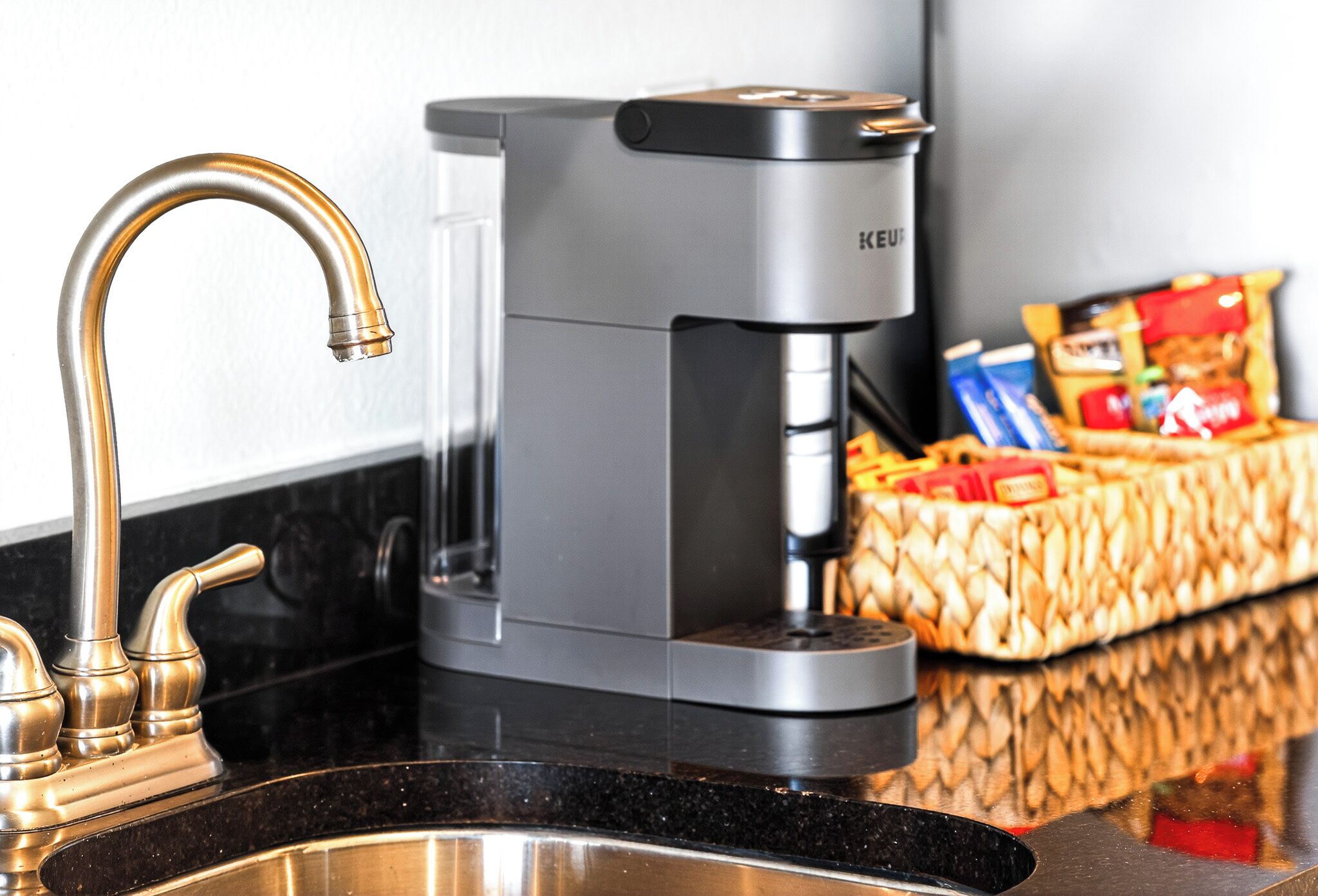 A kitchen with a coffee maker, a sink, and a basket of snacks.
