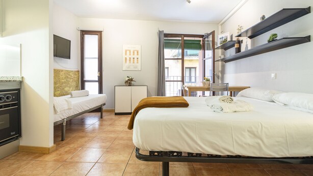 A compact hotel room with a bed, a small desk, a chair, a TV, a kitchenette, and a window with a view of buildings.