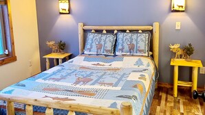 Un dormitorio acogedor con cabecera de cama de madera, colcha con estampados de animales, dos mesitas de noche con lámparas y una decoración de alce montada en la pared.
