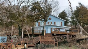 A two-story house with a blue exterior and a star on the roof, surrounded by trees and a dock extending into a body of water. - Lake House with Multiple Decks and Large Swim Platform on Private Dock, (Rocky Mount)