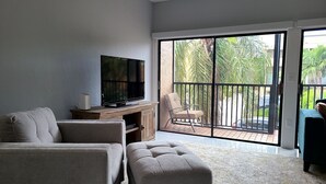 A living room with a flat-screen TV on a wooden cabinet, a grey sofa, a matching ottoman, and a balcony with a chair.