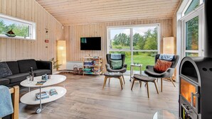 A cozy living room with a fireplace, wooden walls, a flat-screen TV, and a view of the garden. - Cozy home in ålbæk with kitchen (Ålbæk)
