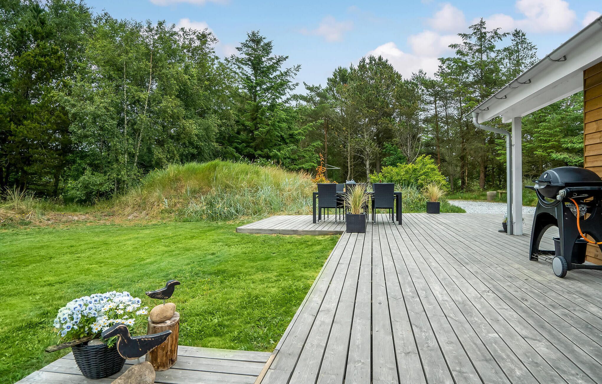 Um deck de madeira com móveis para área externa, churrasqueira e um jardim com uma escultura de pássaro.