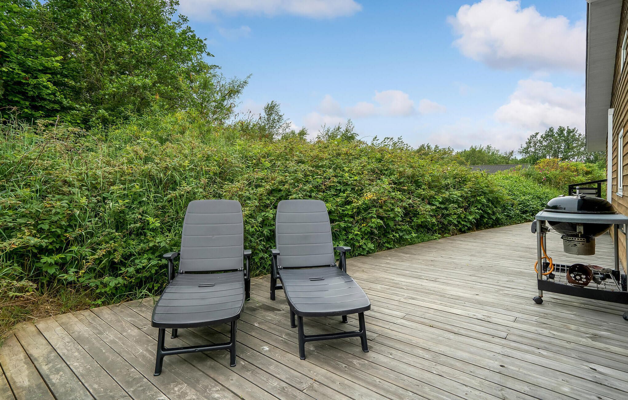 Zwei schwarze Lounge-Stühle auf einer Holzterrasse mit Grill und Grillutensilien zur Rechten.