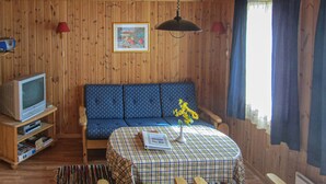 L’intérieur d’un chalet en bois, avec un canapé bleu, un téléviseur posé sur un meuble en bois, une table recouverte d’une nappe à carreaux et une fenêtre avec des rideaux.