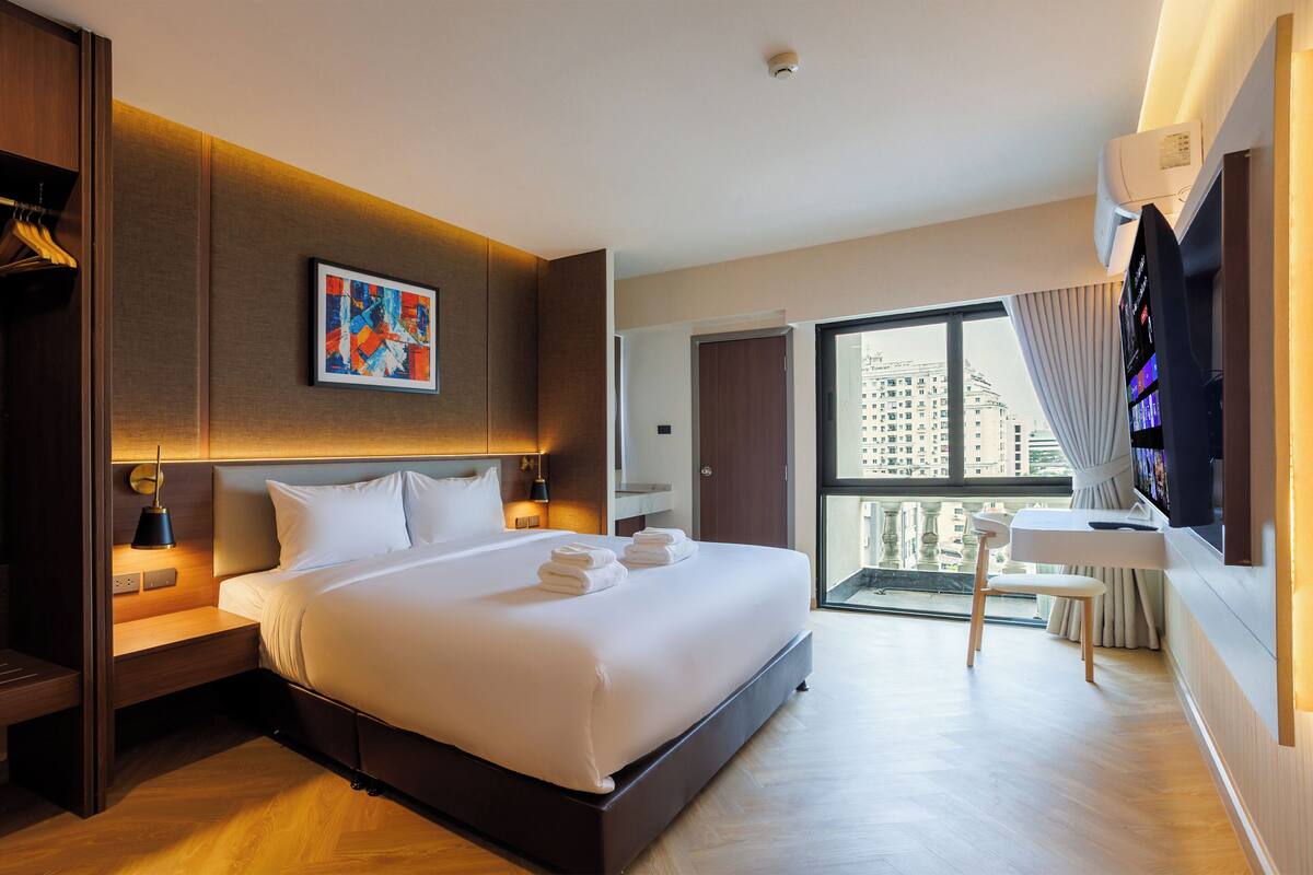 Una habitación de hotel moderna con una cama grande, un escritorio, una silla, un televisor y un balcón con vistas a la ciudad.
