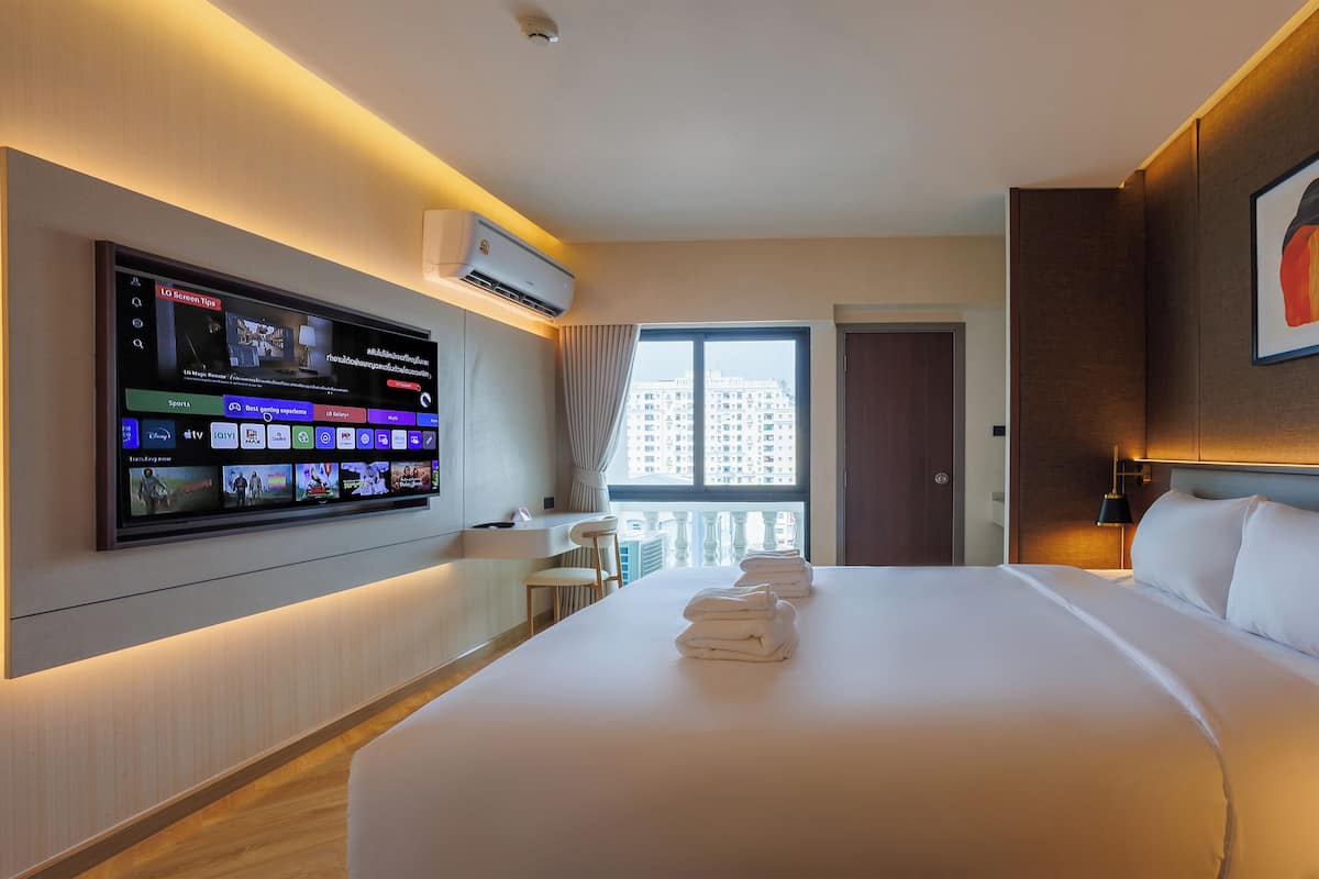 Una habitación de hotel con una cama grande, un televisor de pantalla plana montado en la pared, una ventana con cortinas y una obra de arte enmarcada sobre la cama.