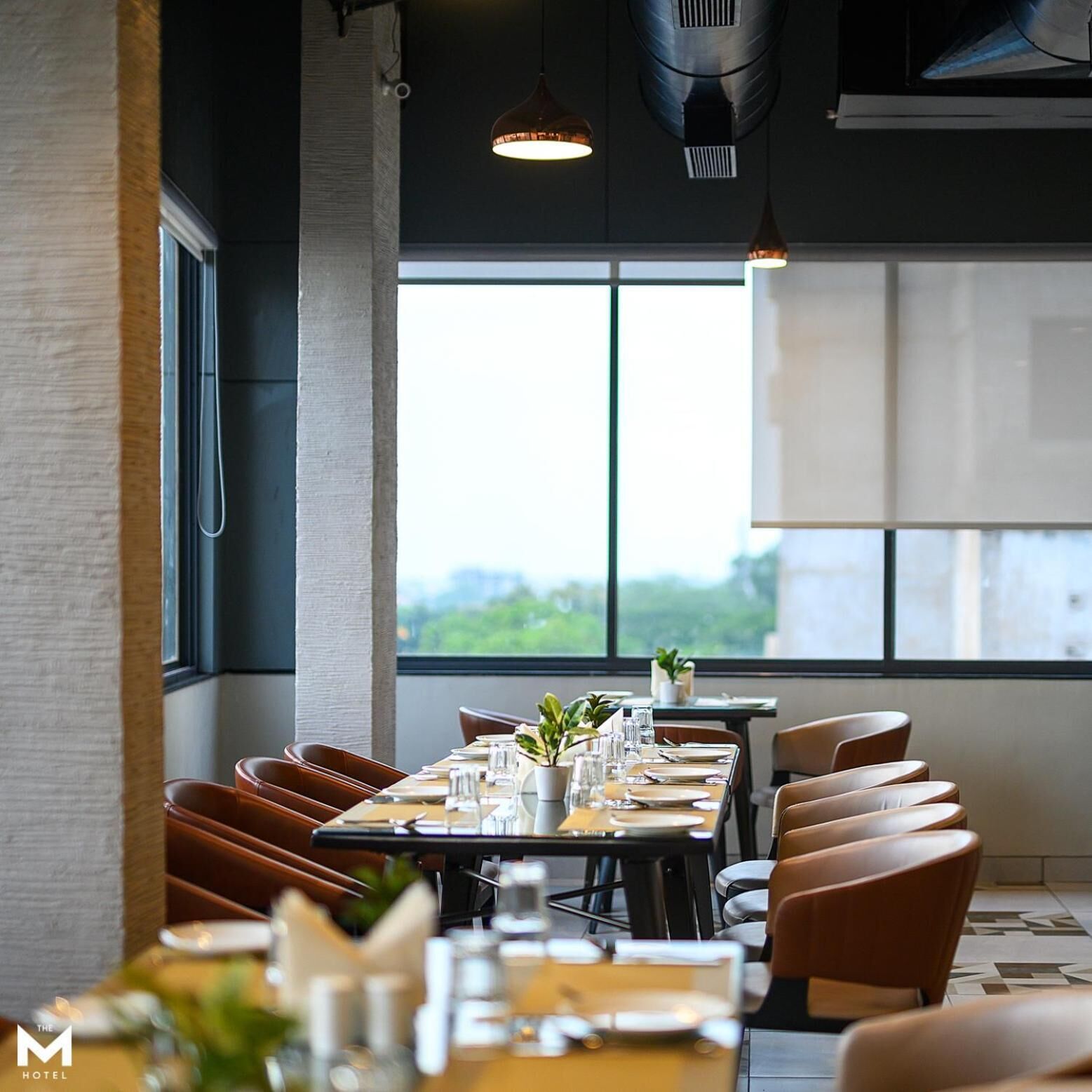 A restaurant with a long table set for dining, large windows, and hanging pendant lights.