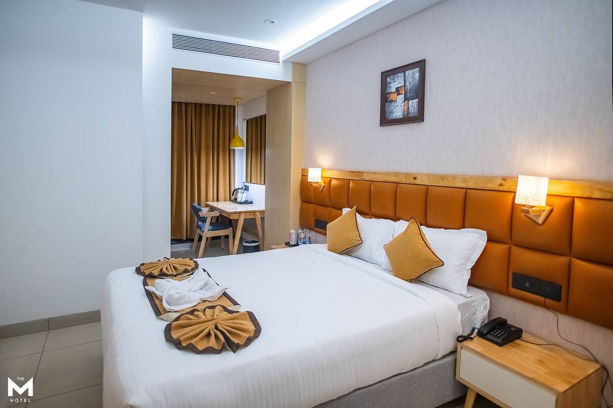 A hotel room with a neatly made bed, a bedside table with a lamp, a desk with a chair, and a window with curtains.