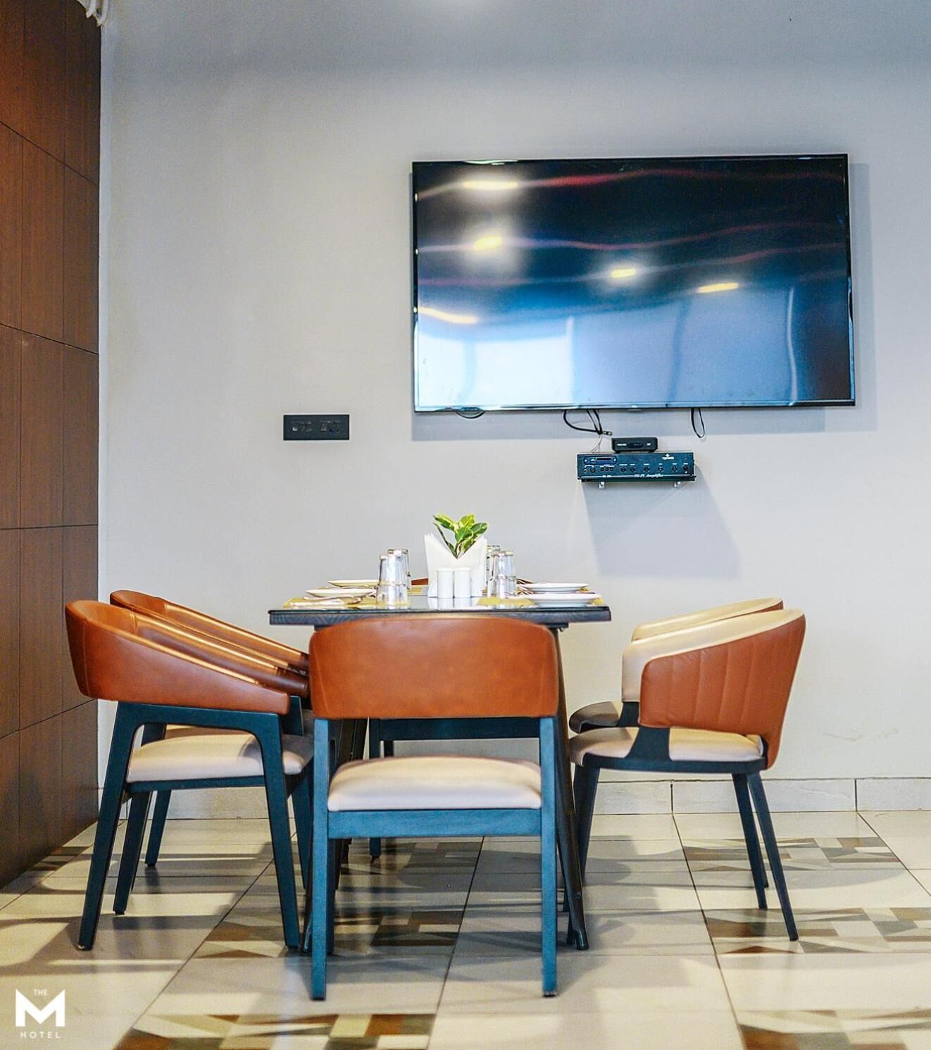 Un comedor moderno con un televisor de pantalla plana grande montado en la pared, una mesa redonda con sillas y suelo de baldosas.