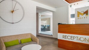 Ett väntrum med en receptionsdisk märkt med "RECEPTION", ett runt vitt bord, en beige soffa med gröna kuddar och en väggklocka.