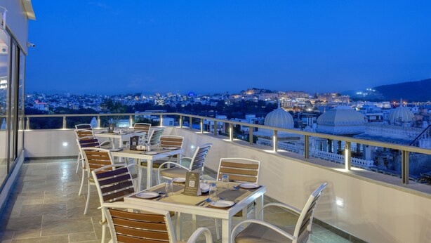 A rooftop terrace with tables and chairs, overlooking a cityscape and a dome-shaped building. - Dev Panache (Udaipur)