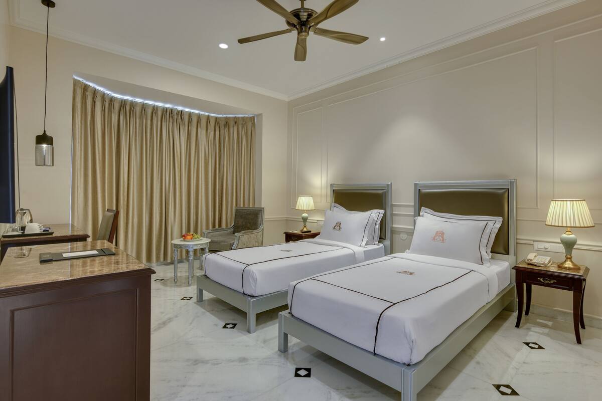 Habitación de hotel con dos camas, un escritorio, una silla, un ventilador de techo y una ventana con cortinas.