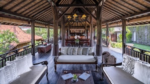 A covered outdoor seating area with a wooden bench and chairs, a table with a plant, and a view of a garden and buildings in the background. - Villa Kapungkur (Jimbaran)