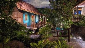 A traditional wooden house with a thatched roof, surrounded by lush tropical vegetation and a stone wall. - Villa Kapungkur (Jimbaran)