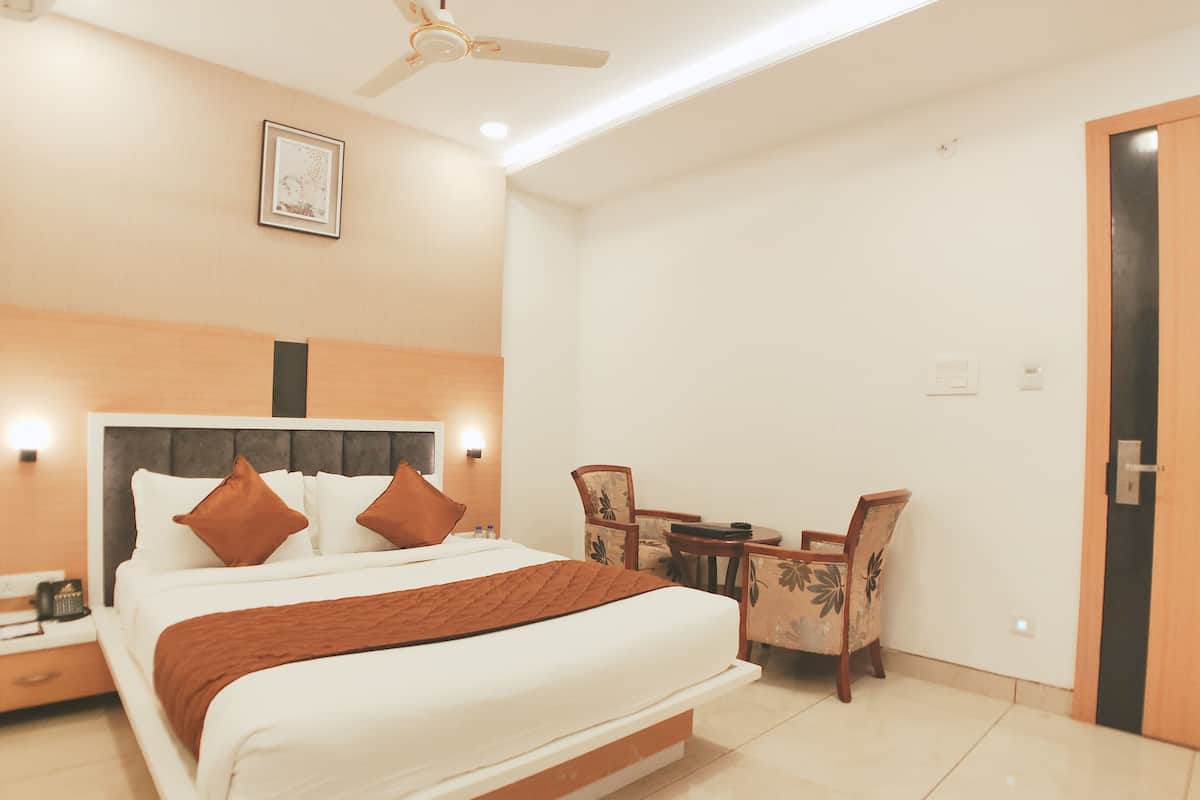 A hotel room with a bed, bedside table, nightstand, a ceiling fan, a wall-mounted light, a framed picture, a dining table with chairs, and a door.