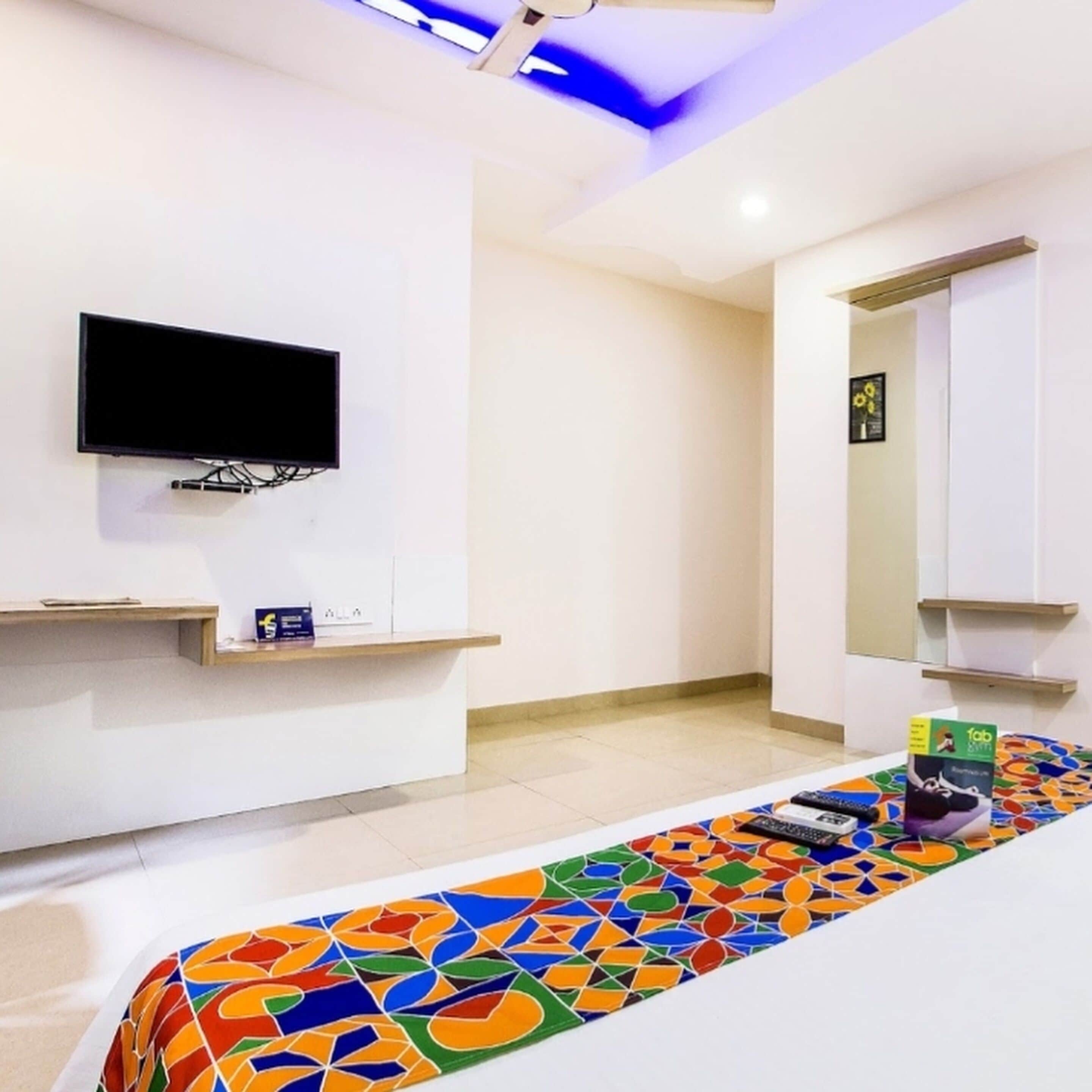 A hotel room with a flat-screen TV mounted on the wall, a small shelf with a few items, a colorful rug, and a bed with a patterned bedspread.