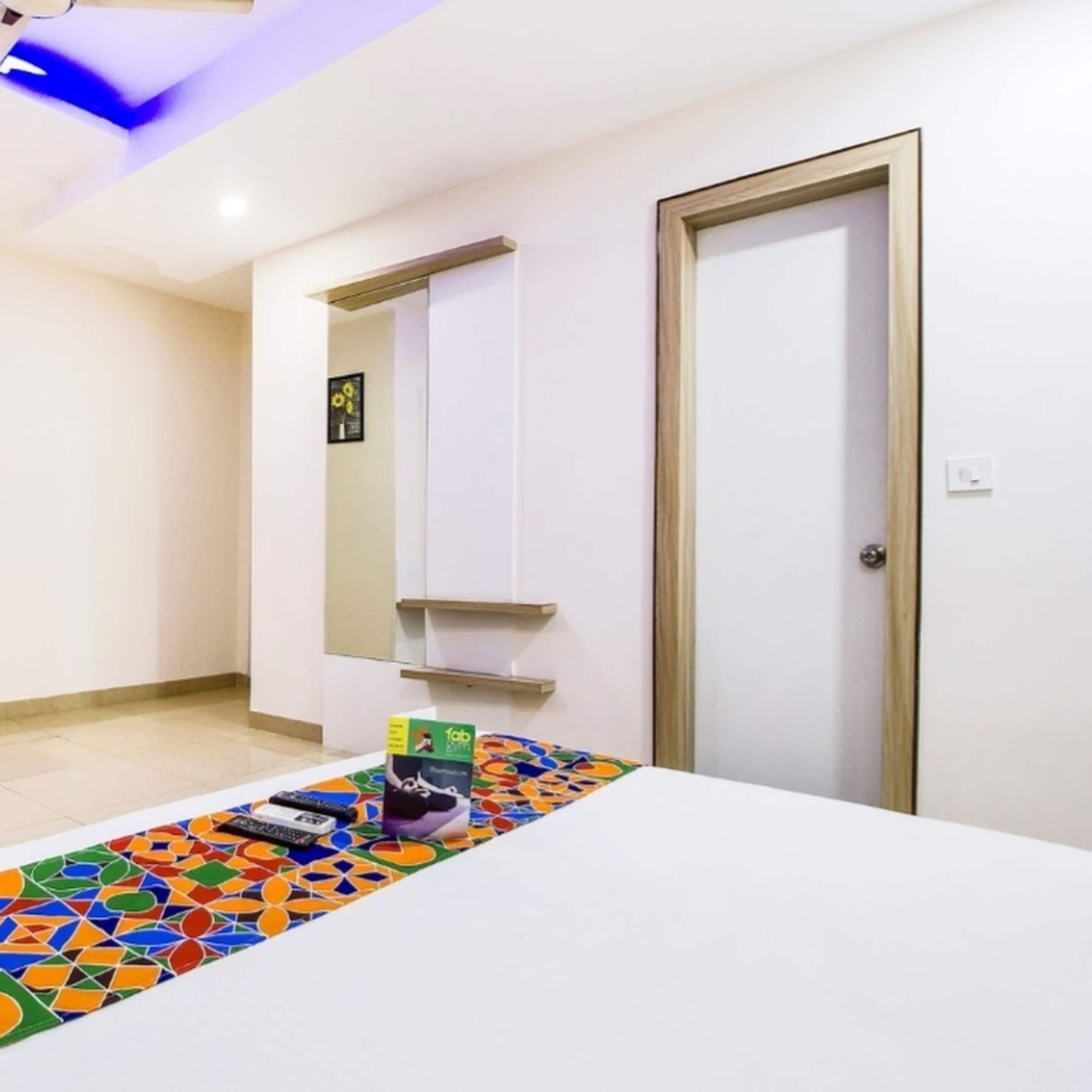 Quarto de hotel com uma cama, um tapete colorido, uma mesa pequena com um controle remoto e uma porta com um espelho.