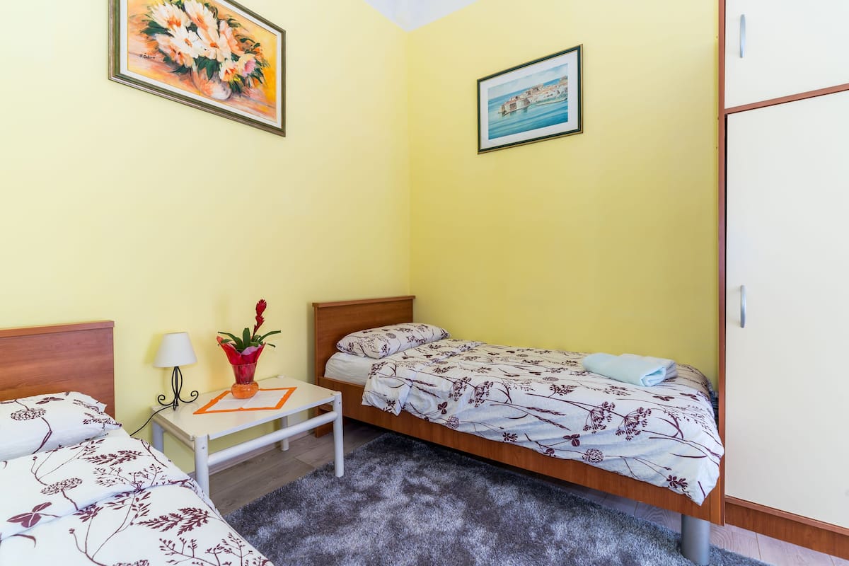 A room with two beds, a small table with a lamp, and a vase with flowers.