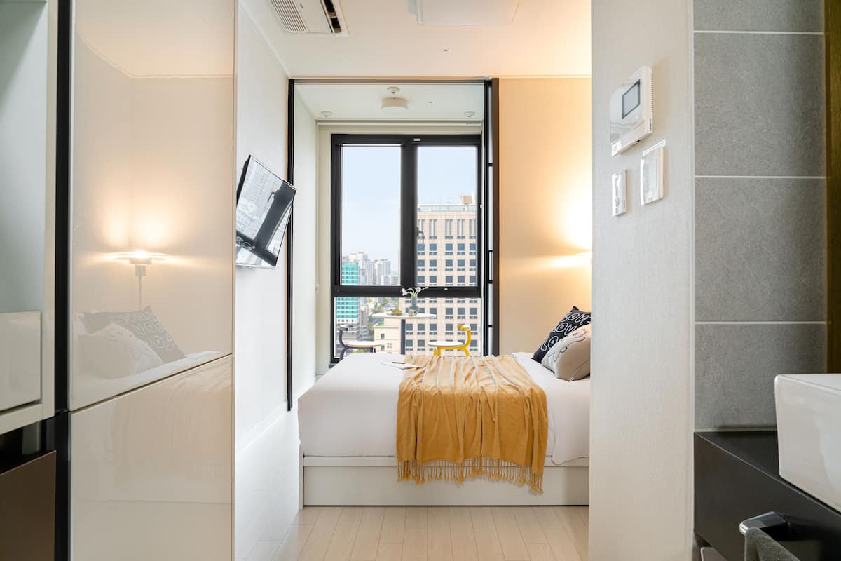 A modern bedroom with a large window, a bed with a yellow throw, and a city view.