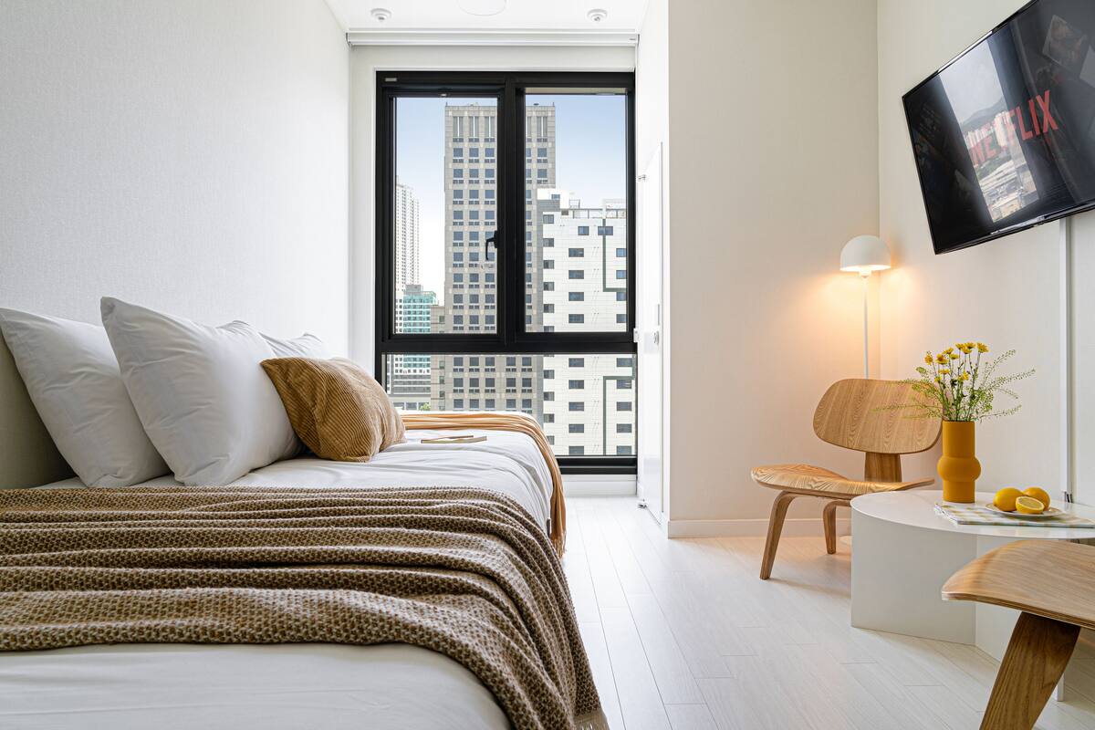 A modern hotel room with a large bed, a desk, a chair, a TV, and a window with a city view.