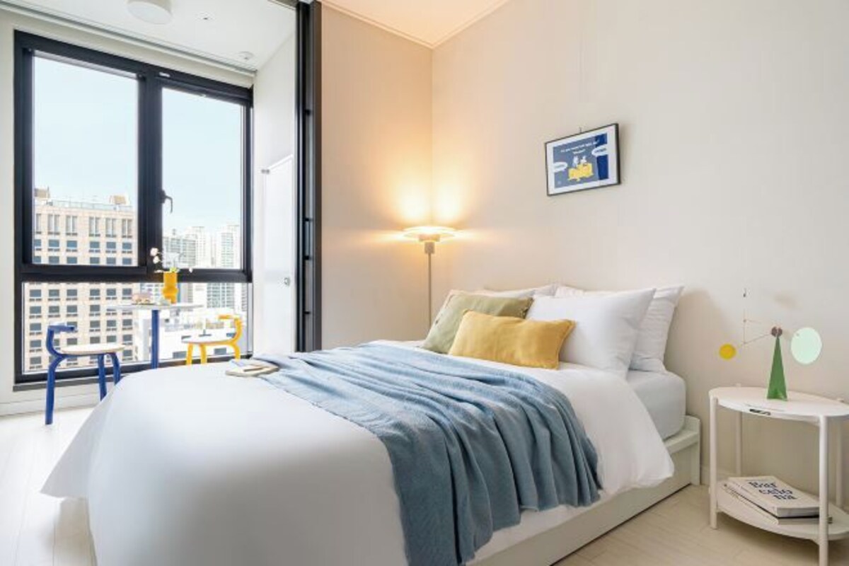 A modern bedroom with a large bed, a bedside table, a nightstand, a window with a view of buildings, and a wall-mounted light fixture.