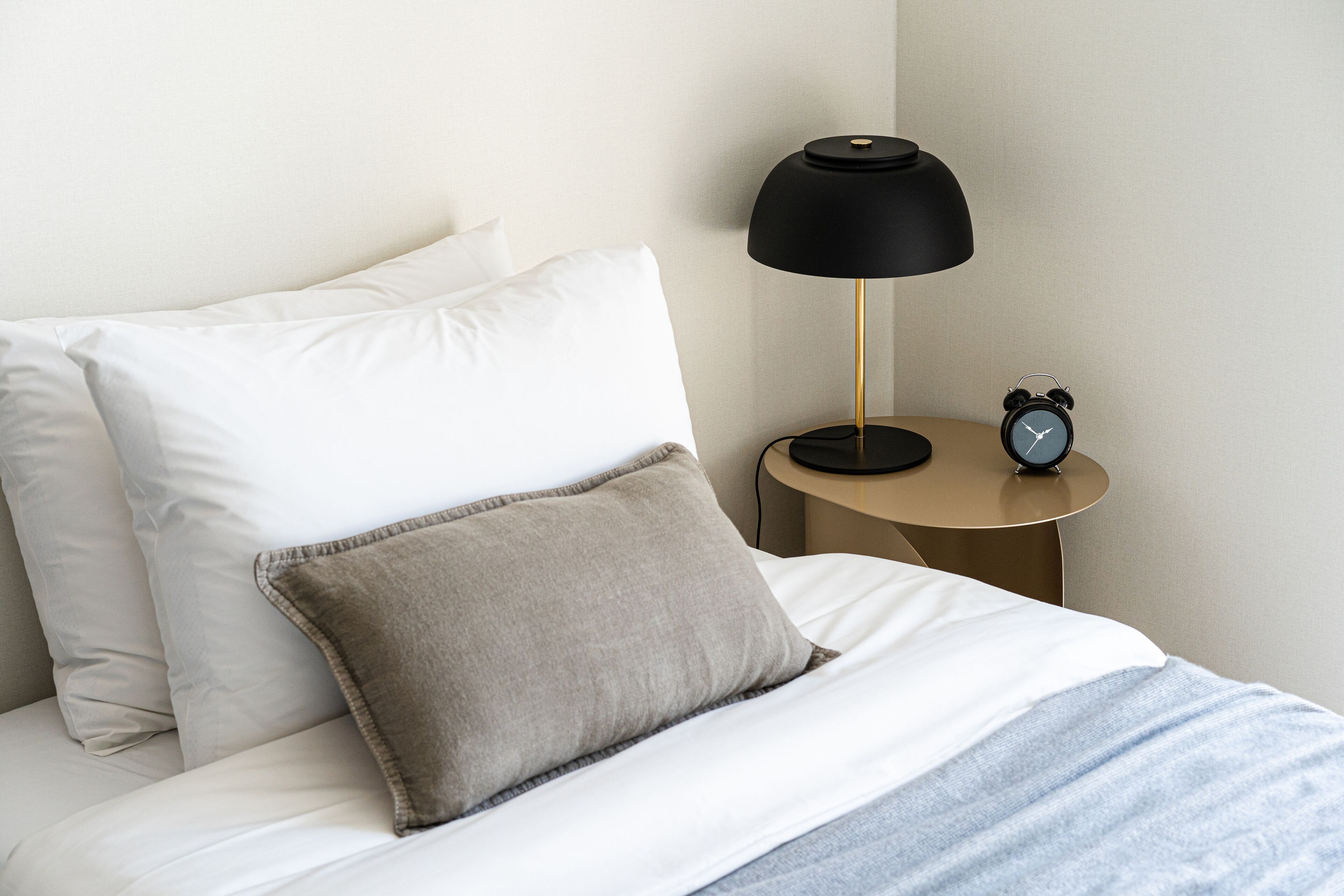 A neatly made bed with white and beige pillows, a black lamp, and a small table with an alarm clock.