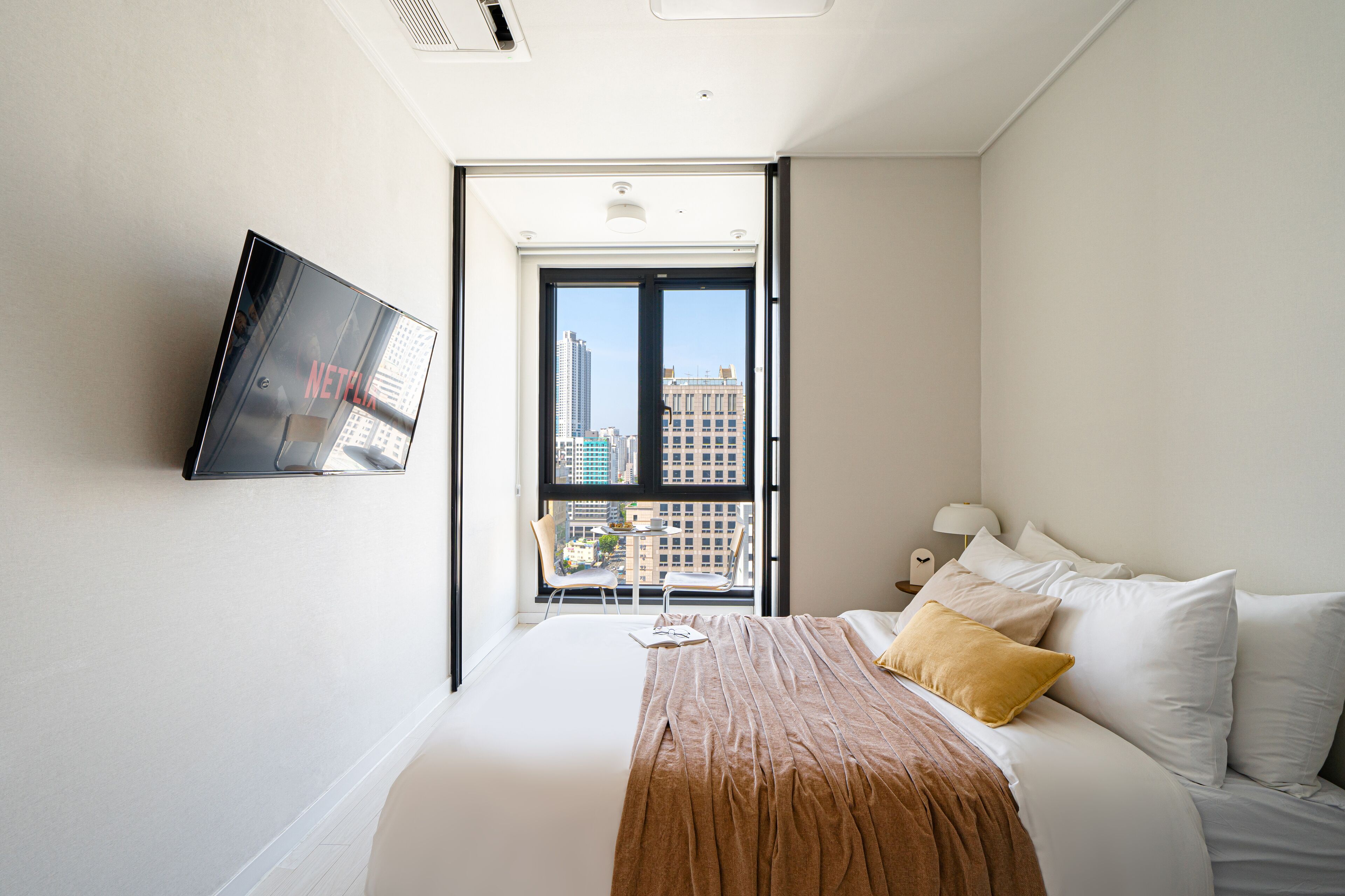 A hotel room with a bed, a TV mounted on the wall, and a window with a city view.