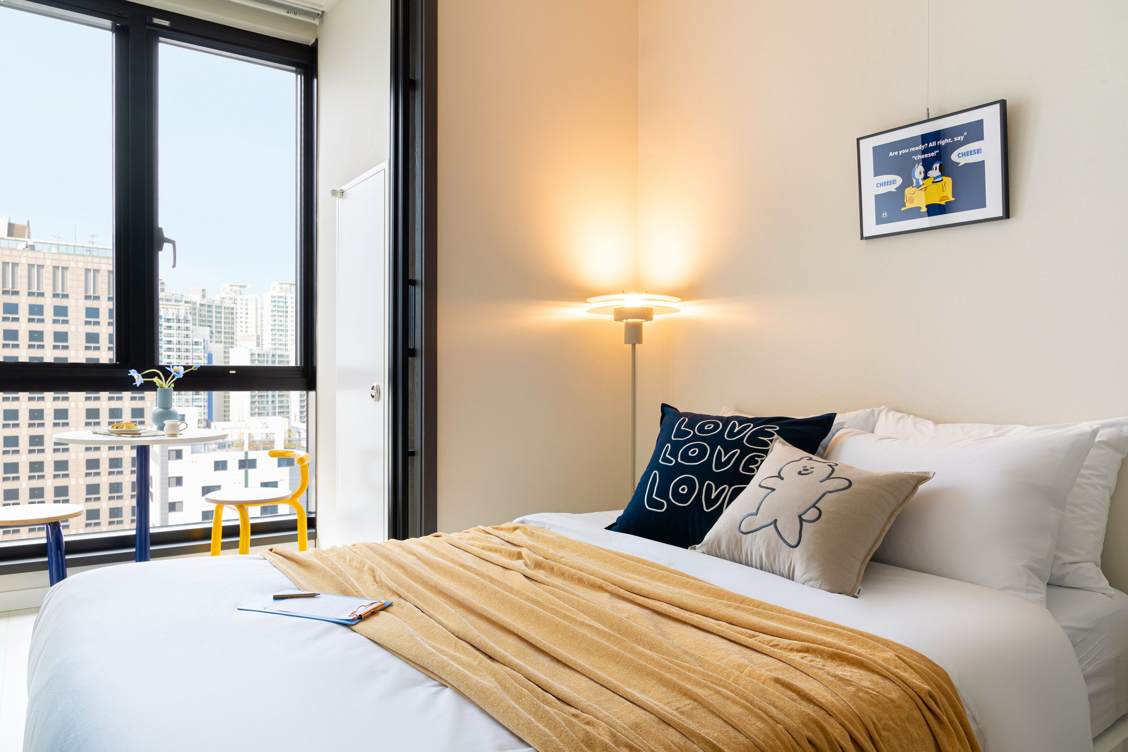 A hotel room with a large window, a bed with white bedding, a yellow throw blanket, a wall-mounted lamp, and a framed picture on the wall.