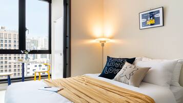 A hotel room with a large window, a bed with white bedding, a yellow throw blanket, a wall-mounted lamp, and a framed picture on the wall.