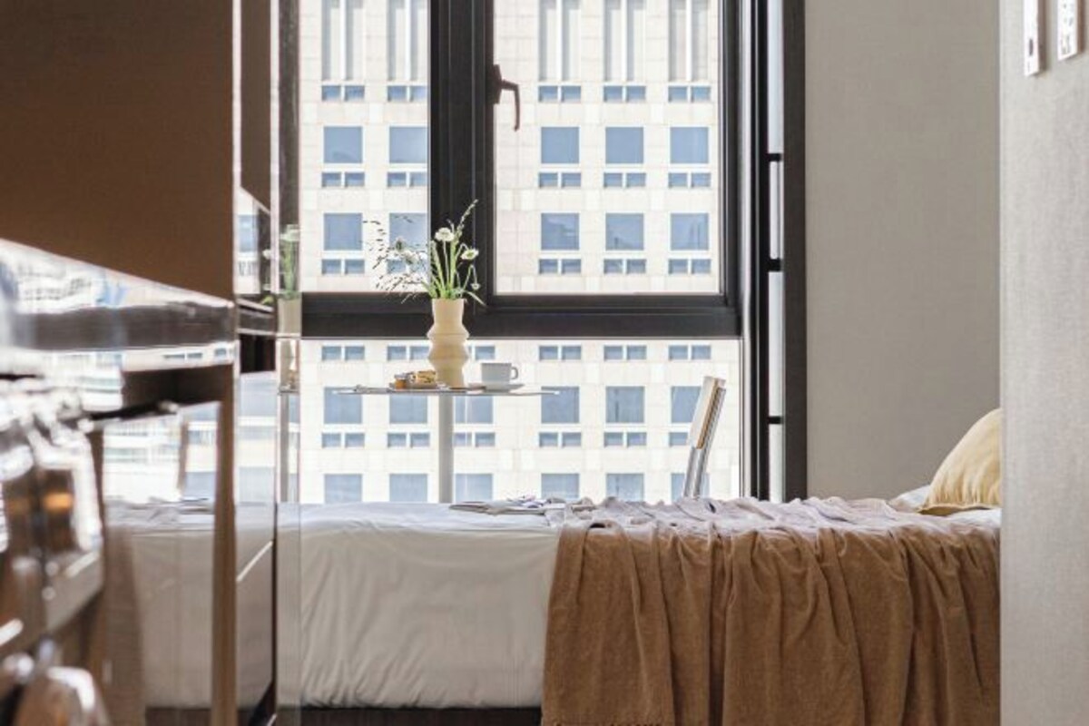 A hotel room with a bed, a bedside table with a vase of flowers, and a window with a view of a building.