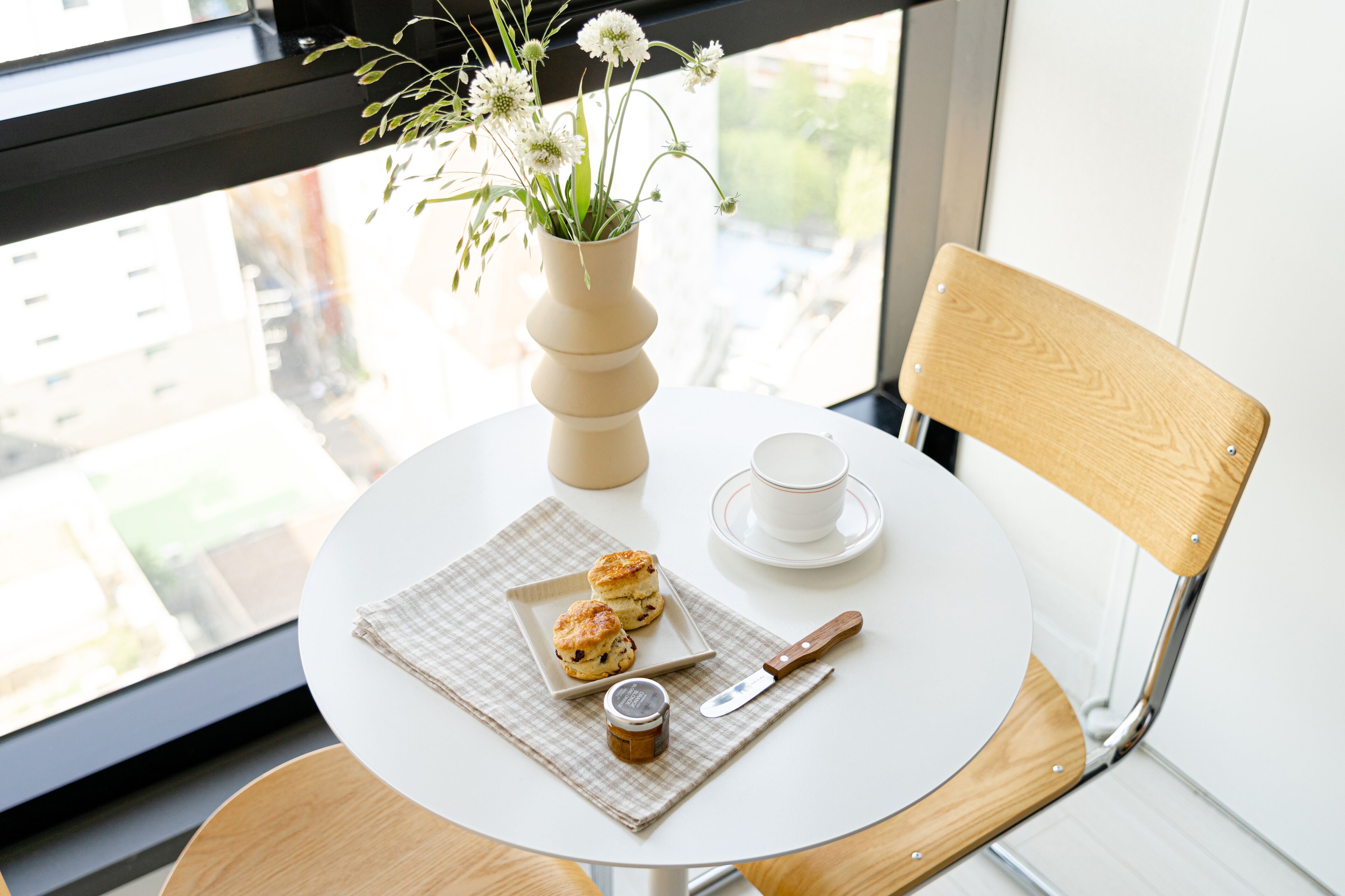 A round table with a white cup, saucer, two scones, a knife, a jar of jam, and a vase with flowers.