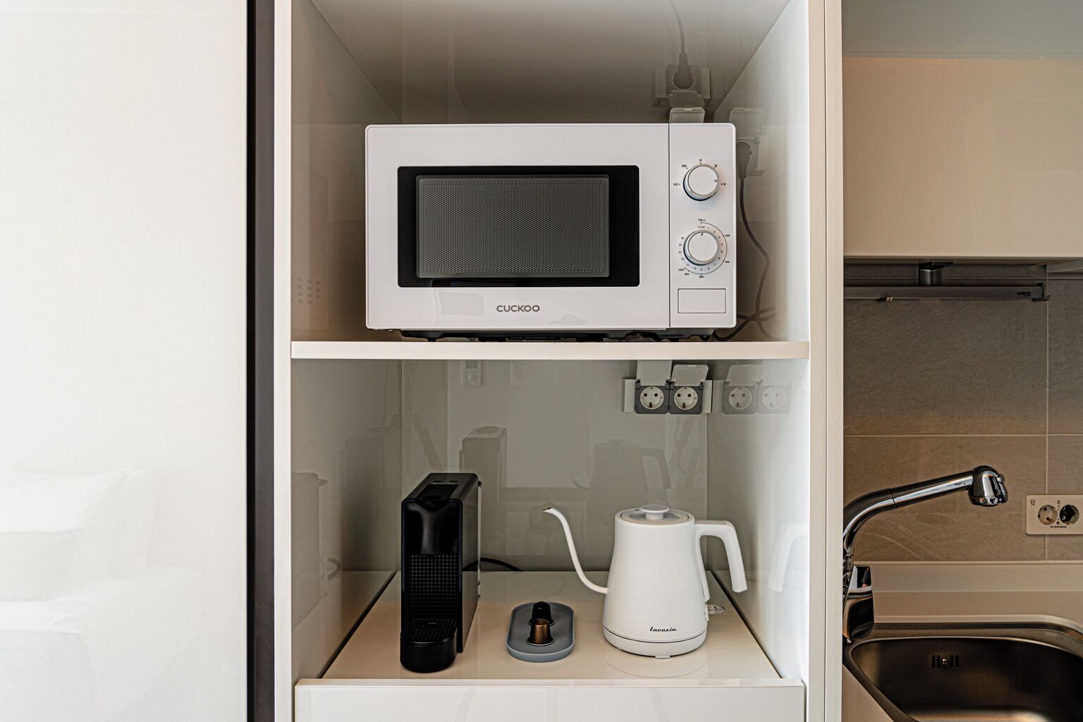 Une petite cuisine équipée d’un four à micro-ondes, d’une machine à café et d’une bouilloire, le tout posé sur un plan de travail blanc.