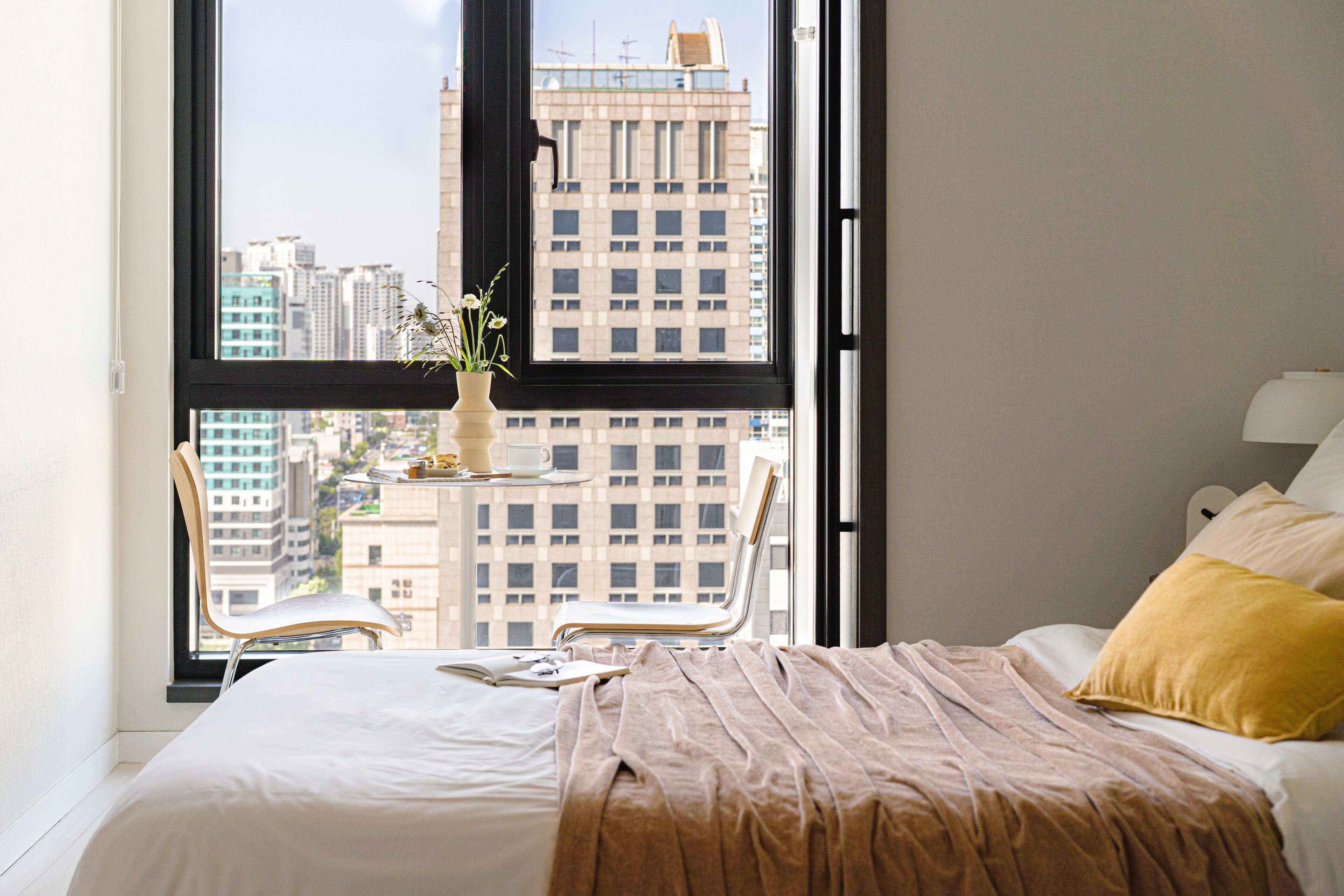 A bedroom with a large window, a bed with a beige blanket, a yellow pillow, and a chair.