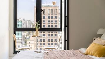A bedroom with a large window, a bed with a beige blanket, a yellow pillow, and a chair.