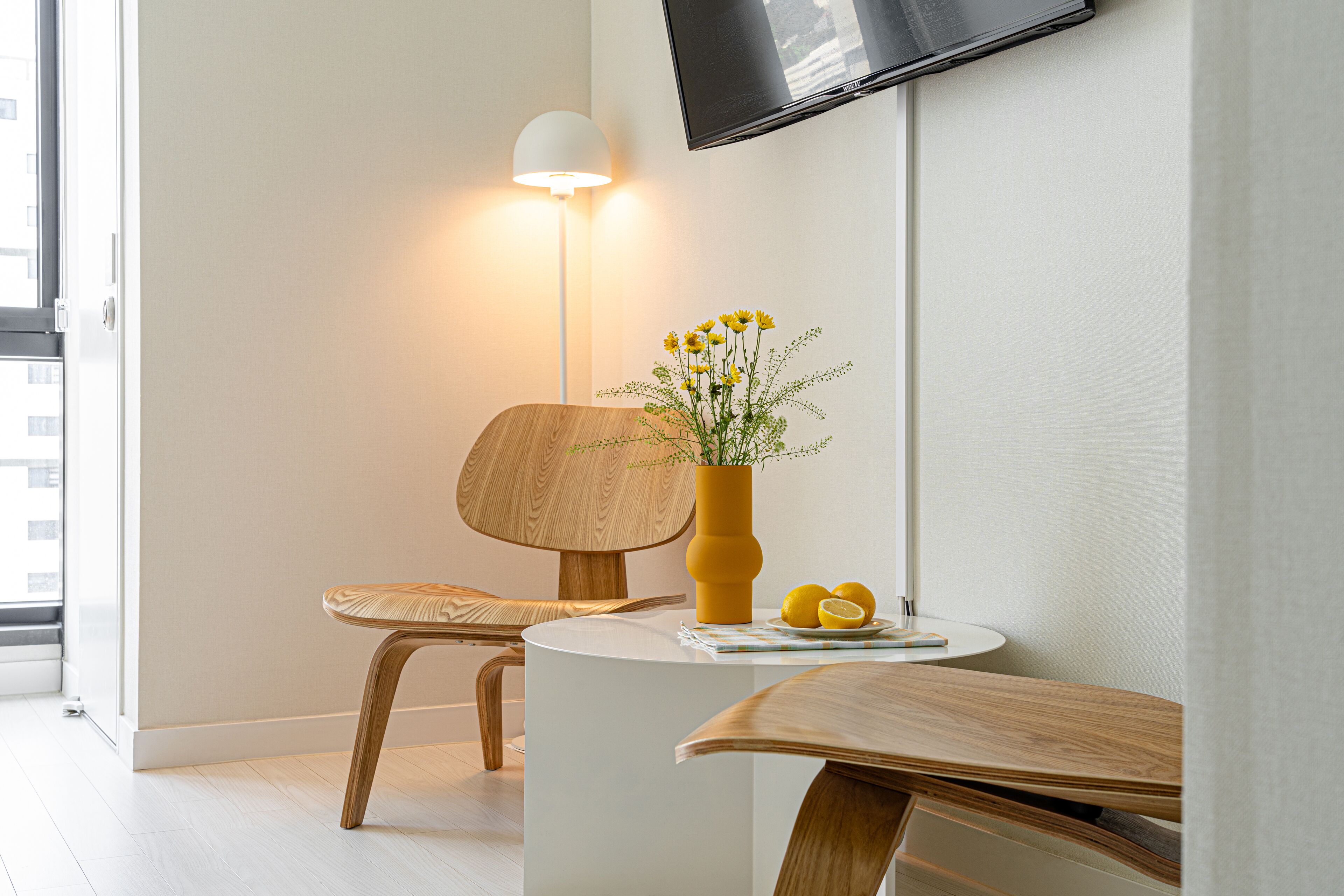 Une pièce moderne avec une table ronde blanche, une chaise en bois, un vase jaune orné de fleurs et un téléviseur à écran plat fixé au mur.