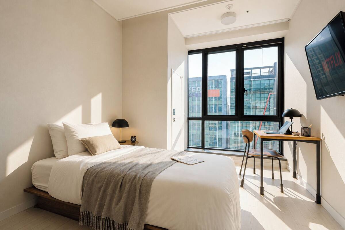 A bedroom with a bed, a desk, a chair, a TV, and a large window with a city view.