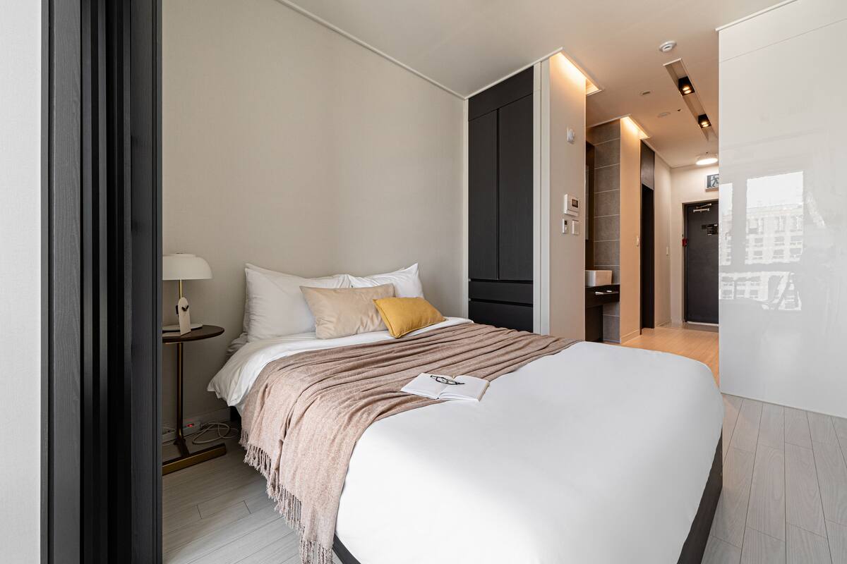 A modern hotel room with a large bed, bedside table, lamp, and a view of the city through the window.