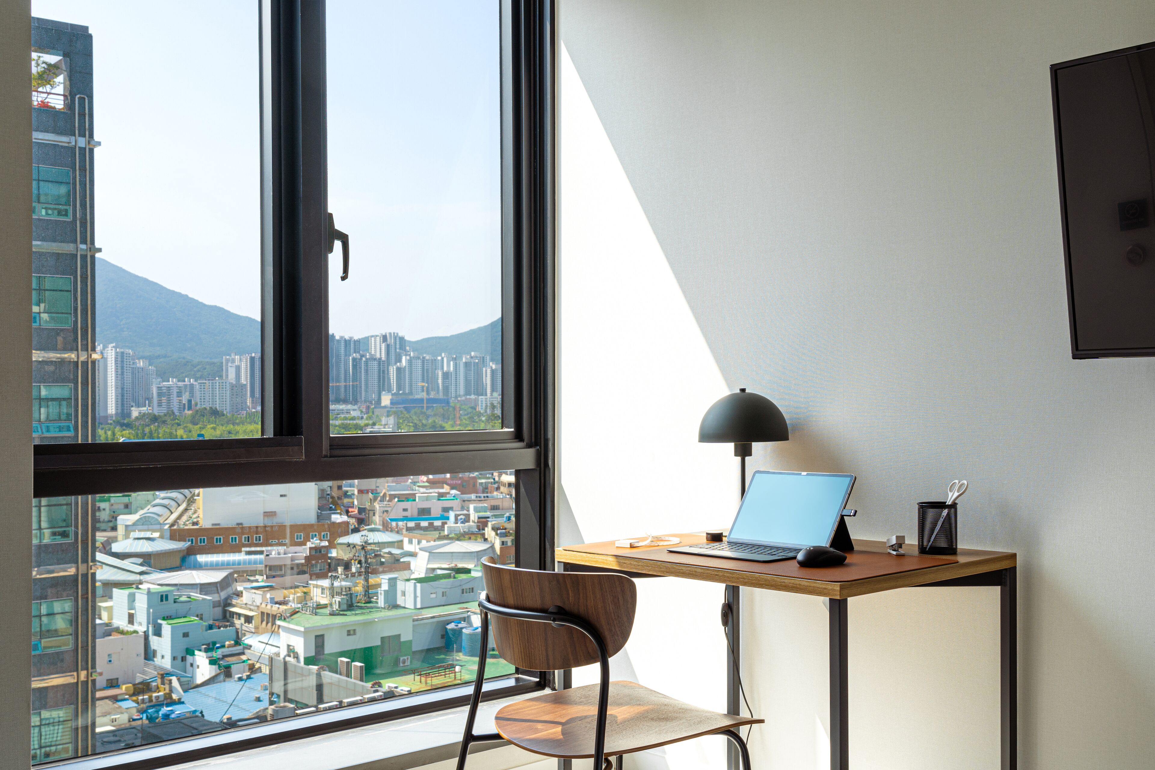 Un espace de bureau moderne comprenant un bureau, une chaise et un ordinateur portable, avec vue sur un paysage urbain composé d’immeubles et d’une montagne.